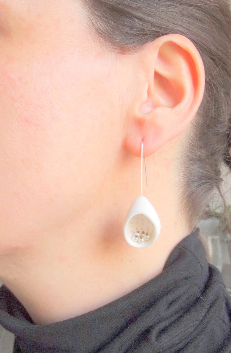 May include: White ceramic earrings with a teardrop shape and small white beads inside. The earrings are hanging from silver hooks.