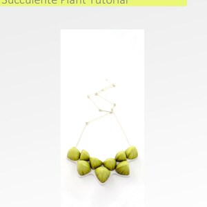 May include: A green succulent plant necklace with a silver chain. The necklace is made of several small, green succulent plants that are arranged in a circle. The chain is attached to the back of the necklace.