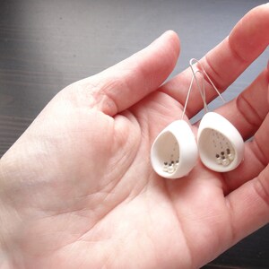 May include: A pair of white ceramic earrings with small white beads inside. The earrings are shaped like a teardrop and have a silver hook.
