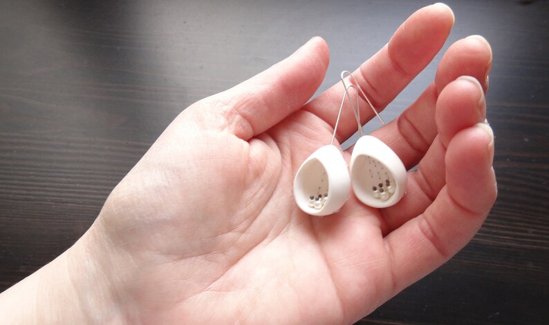 May include: A pair of white ceramic earrings with small white beads inside. The earrings are shaped like a teardrop and have a silver hook.