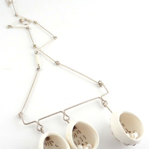 May include: A silver chain necklace with three white ceramic pendants, each containing small white pearls. The pendants are shaped like open egg shells.