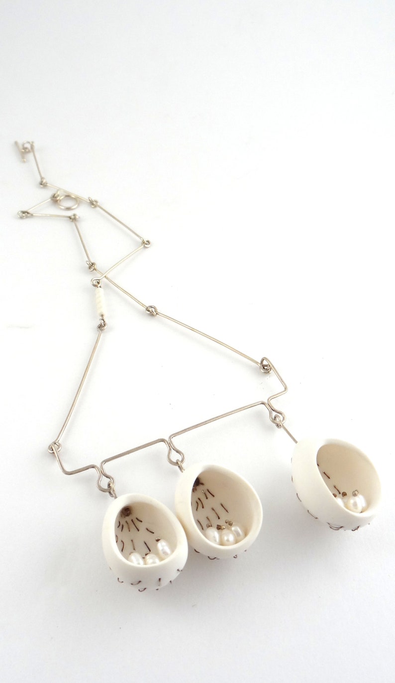 May include: A silver chain necklace with three white ceramic pendants, each containing small white pearls. The pendants are shaped like open egg shells.