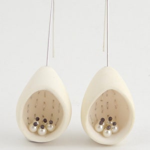 May include: A pair of white ceramic earrings with a teardrop shape. Each earring has a small opening with a cluster of white pearls and black beads inside.