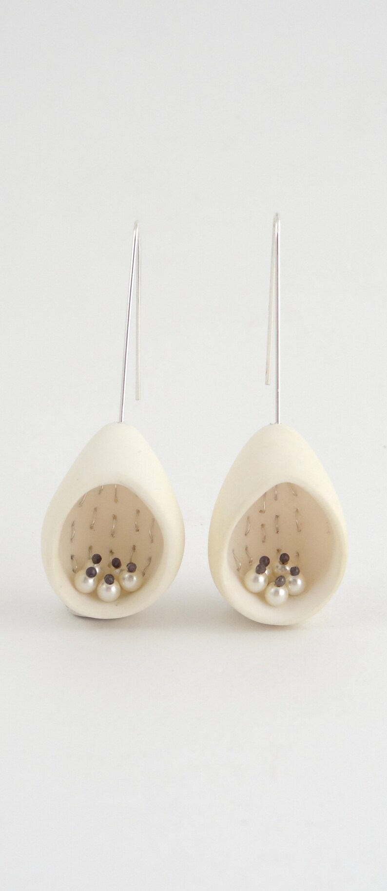 May include: A pair of white ceramic earrings with a teardrop shape. Each earring has a small opening with a cluster of white pearls and black beads inside.