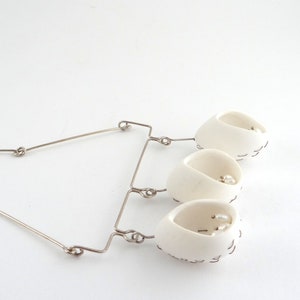 May include: A white ceramic necklace with three pendant cups, each containing a pearl. The cups are connected by a silver wire.