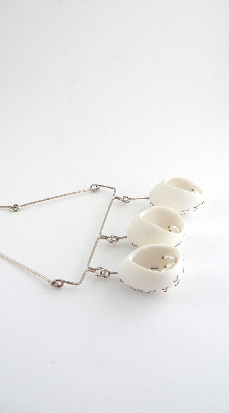 May include: A white ceramic necklace with three pendant cups, each containing a pearl. The cups are connected by a silver wire.