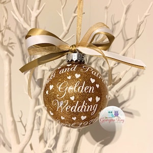 May include: Gold glitter Christmas ornament with white text that reads "[Name] and [Name] Golden Wedding April 1970". The ornament is decorated with white hearts and has a gold and white ribbon.