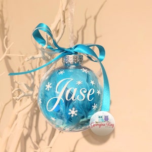 May include: A clear glass Christmas bauble with a blue background and white snowflakes. The name "Jase" is written in white on the bauble. The bauble is decorated with white feathers and a blue ribbon.