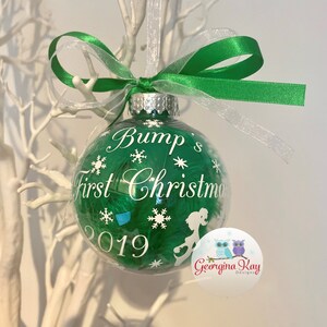May include: A clear Christmas bauble filled with green feathers, featuring the text "Bump's First Christmas 2019". It is adorned with white snowflakes and a green and white ribbon for hanging.