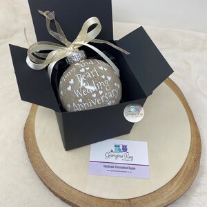 May include: A beige glass ornament with white text that reads "Mum and Dad Pearl Wedding Anniversary" and white hearts. The ornament is in a black gift box with a gold ribbon. The box is on a wooden surface.