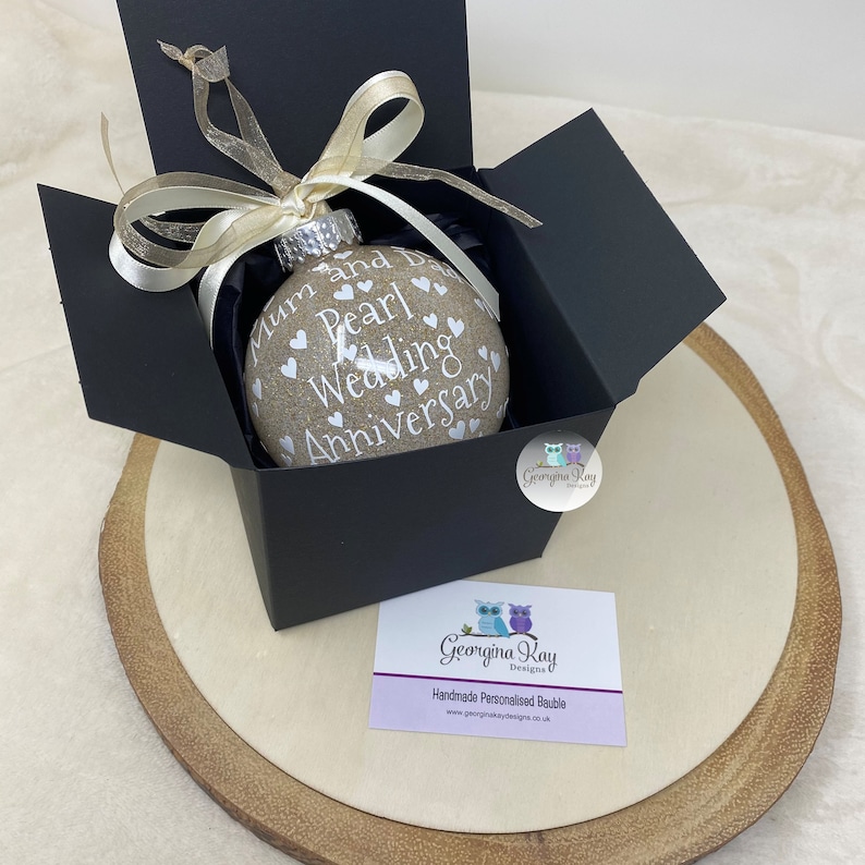 May include: A beige glass ornament with white text that reads "Mum and Dad Pearl Wedding Anniversary" and white hearts. The ornament is in a black gift box with a gold ribbon. The box is on a wooden surface.