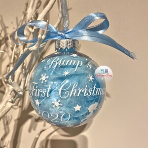 May include: A clear bauble filled with blue feathers, featuring white snowflakes and stars. The text reads "Bump's First Christmas 2020". A light blue ribbon is tied at the top. A festive decoration.