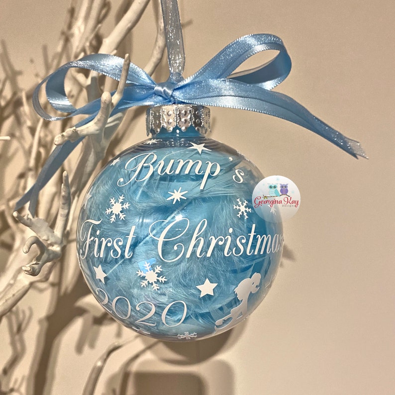 May include: A clear bauble filled with blue feathers, featuring white snowflakes and stars. The text reads "Bump's First Christmas 2020". A light blue ribbon is tied at the top. A festive decoration.