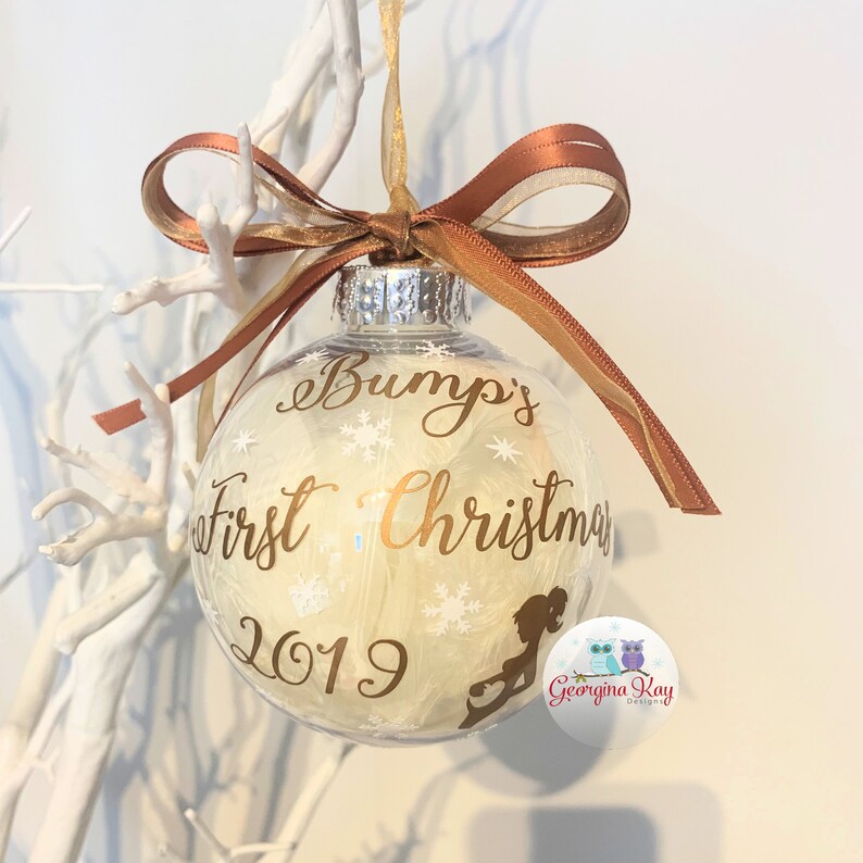 May include: A clear Christmas bauble with gold text reading "Bump's First Christmas 2019". The bauble is filled with white feathers and snowflakes. It is decorated with a brown and gold ribbon.