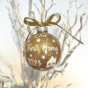 May include: Gold glitter Christmas ornament with the text "Our First Home 2023" and a white silhouette of a house. The ornament is hanging from a gold ribbon.