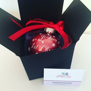 May include: A red glitter Christmas bauble with the text "Joshua's 1st Christmas" and snowflake designs, presented in a black gift box with a red ribbon. A business card is visible.