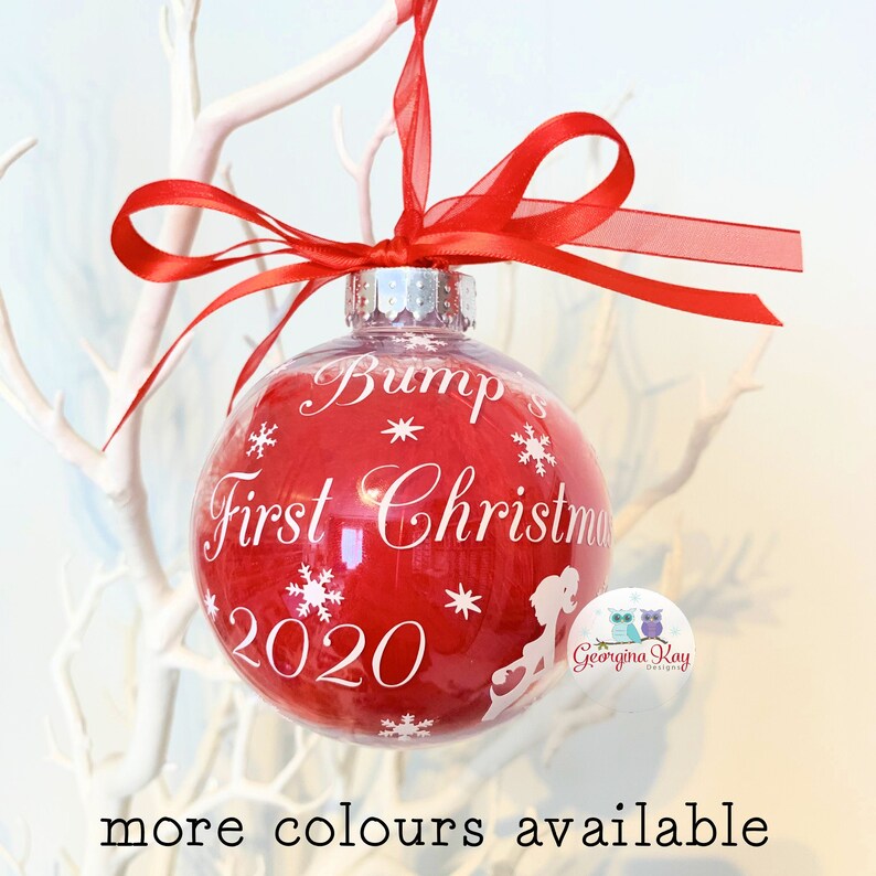 May include: A red Christmas bauble with the text "Bump's First Christmas 2020" in white. The bauble features snowflake and silhouette designs and is tied with a red ribbon. It hangs from a white branch.