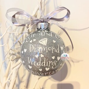 Puede incluir: Un adorno navide&ntilde;o de purpurina plateada con un diamante blanco y el texto "Maggie and Gary Diamond Wedding Anniversary".