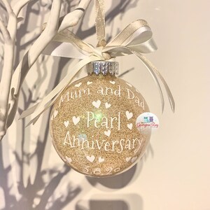 May include: Gold glitter Christmas ornament with white text that reads "Mum and Dad Pearl Anniversary 3.8.20". The ornament is decorated with white hearts.
