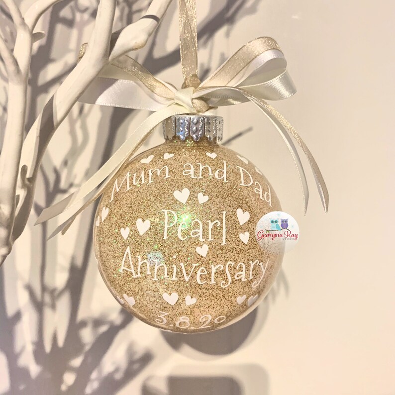 May include: Gold glitter Christmas ornament with white text that reads "Mum and Dad Pearl Anniversary 3.8.20". The ornament is decorated with white hearts.