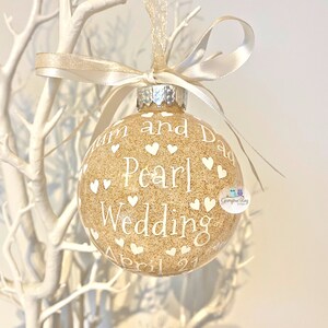 May include: A gold glitter Christmas ornament with white hearts and the text "Mum and Dad Pearl Wedding April 21st".