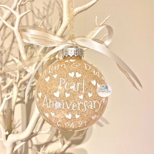May include: A gold glitter Christmas ornament with white text that reads "Mum & Dad Pearl Anniversary 06.04.21". The ornament is decorated with white hearts.