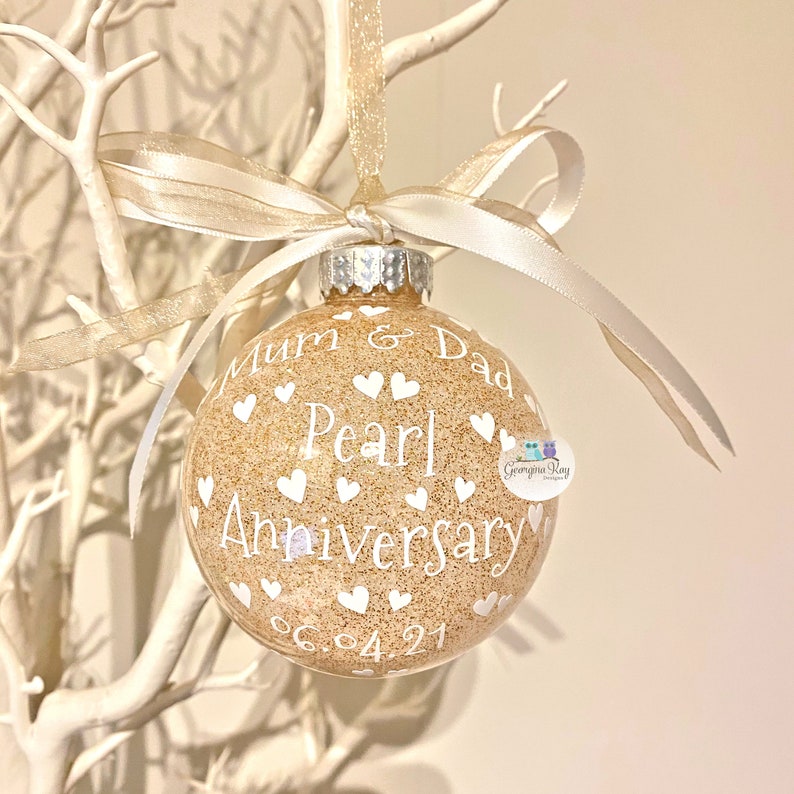 May include: A gold glitter Christmas ornament with white text that reads "Mum & Dad Pearl Anniversary 06.04.21". The ornament is decorated with white hearts.