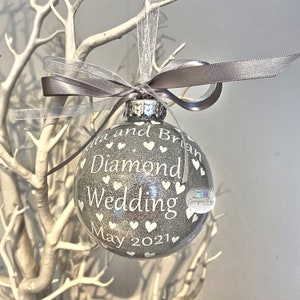 Puede incluir: Un adorno navide&ntilde;o de purpurina plateada con corazones blancos y el texto "Rita and Brian Diamond Wedding May 2021".