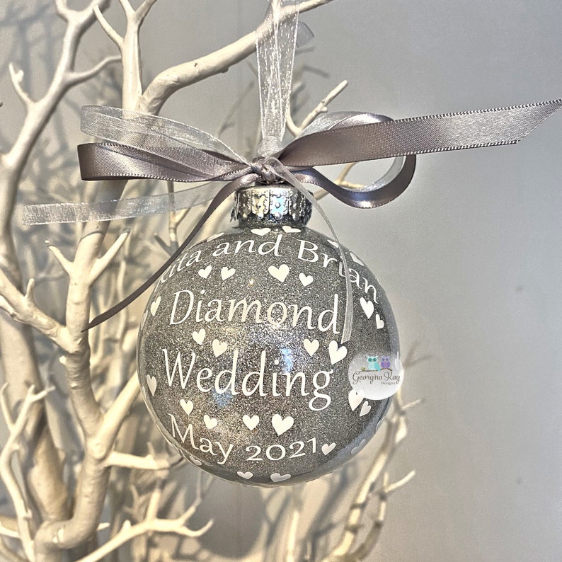Puede incluir: Un adorno navide&ntilde;o de purpurina plateada con corazones blancos y el texto "Rita and Brian Diamond Wedding May 2021".
