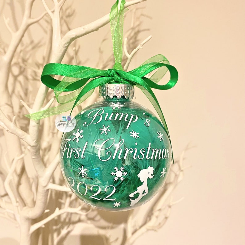 May include: A clear glass Christmas bauble filled with green feathers, featuring white text and snowflake designs. The text reads "Bump's First Christmas 2022" and includes a pregnant person silhouette. A green ribbon is tied at the top.