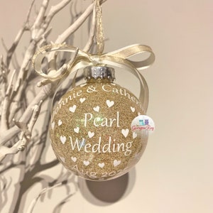 May include: Gold glitter Christmas ornament with white hearts and the text "Ronnie & Catherine Pearl Wedding Aug 2022".