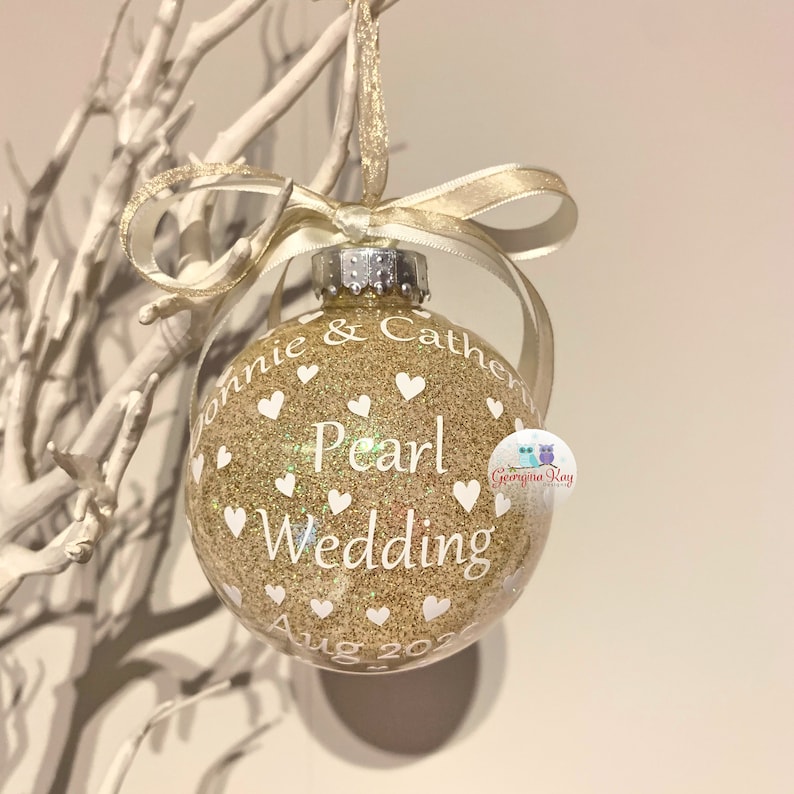 May include: Gold glitter Christmas ornament with white hearts and the text "Ronnie & Catherine Pearl Wedding Aug 2022".