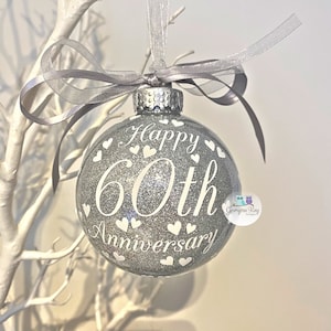 Puede incluir: Adorno navide&ntilde;o de purpurina plateada con texto blanco que dice "Feliz 60 aniversario" y corazones blancos. El adorno est&aacute; colgado de una cinta plateada.