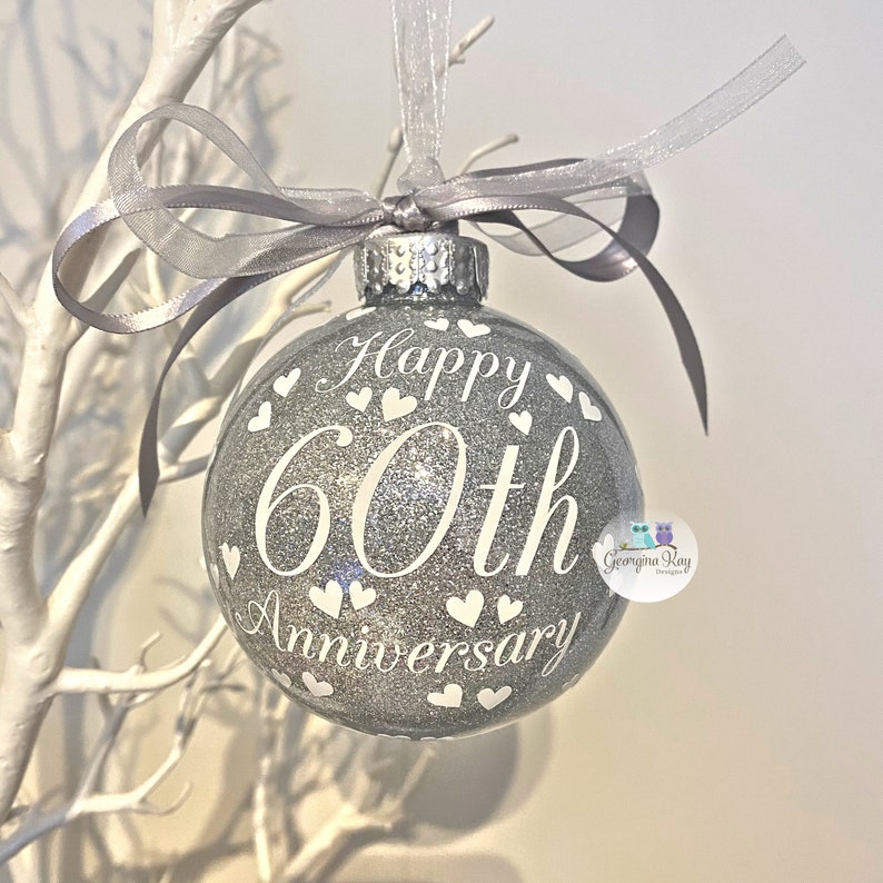 Puede incluir: Adorno navide&ntilde;o de purpurina plateada con texto blanco que dice "Feliz 60 aniversario" y corazones blancos. El adorno est&aacute; colgado de una cinta plateada.