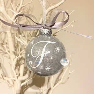 May include: A silver glitter Christmas bauble with a white snowflake design and the letter "F" in a white cursive font. The bauble is hanging from a white ribbon with a silver bow.