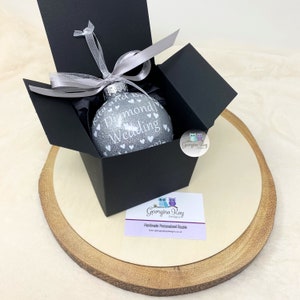 Puede incluir: Un adorno navide&ntilde;o de purpurina plateada con el texto "Diamond Wedding" y peque&ntilde;os corazones blancos. El adorno est&aacute; en una caja de regalo negra con una cinta plateada.