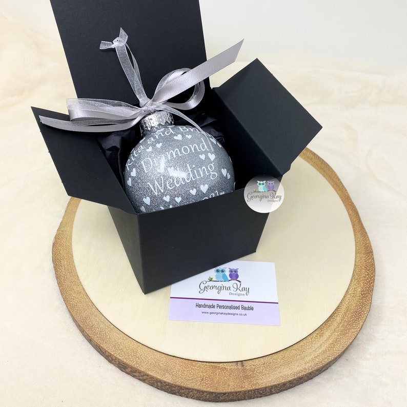 Puede incluir: Un adorno navide&ntilde;o de purpurina plateada con el texto "Diamond Wedding" y peque&ntilde;os corazones blancos. El adorno est&aacute; en una caja de regalo negra con una cinta plateada.