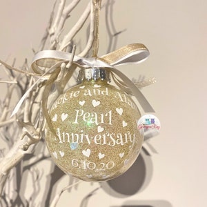 May include: A gold glitter Christmas ornament with white text that reads "Jackie and Alan Pearl 1 Anniversary 6.10.20". The ornament is decorated with white hearts.