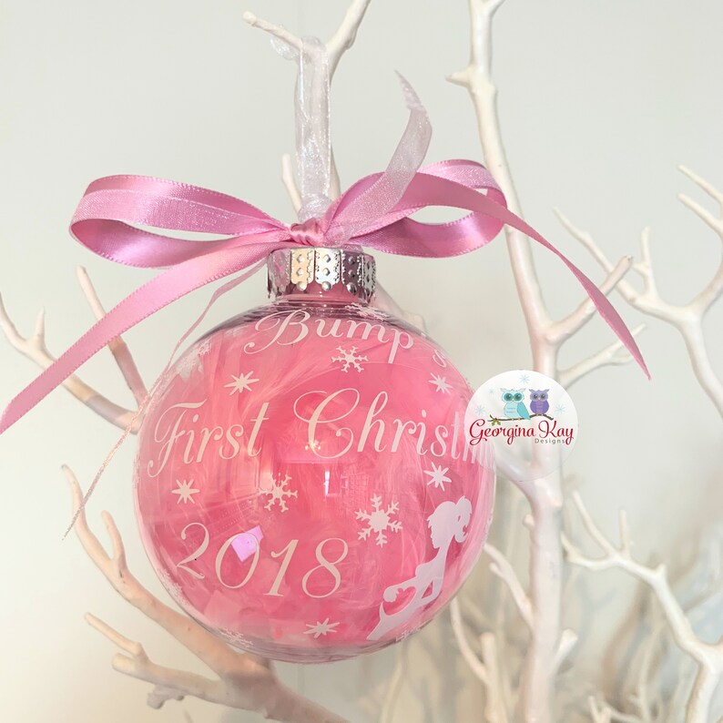 May include: A pink Christmas bauble with the text "Bump First Christmas 2018" and snowflake and silhouette designs. The bauble is filled with pink feathers and has a pink ribbon tied at the top.