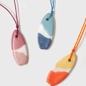 Op de afbeelding: Drie kleurrijke keramische surfplank hangers aan koorden. De hangers zijn geschilderd in tinten blauw, roze en oranje met witte accenten.