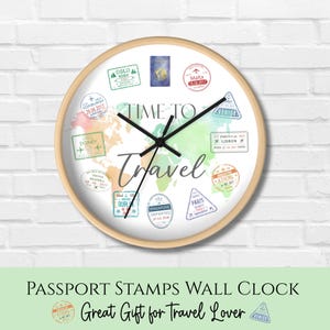 May include: A round wall clock with a light wood frame. The clock face features a world map design with colorful watercolor accents and travel-themed stamps. The words "Time to Travel" are written across the face. The clock hands are black.