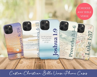 Puede incluir: Cinco fundas para iPhone con versículos bíblicos. Las fundas presentan diseños con temas de atardecer, océano y flores. Los textos incluyen versículos de Romanos, Filipenses, Josué, Proverbios y Lucas. Se muestran sobre una superficie de madera.