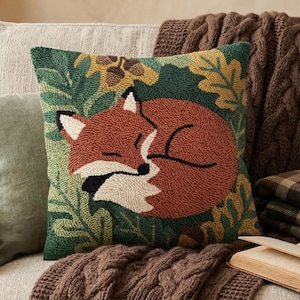 May include: A decorative pillow with a sleeping fox design in shades of orange, brown, and white, set against a green and yellow leaf pattern. The pillow is on a beige sofa, with a brown cable-knit throw, a green pillow, and a plaid blanket.
