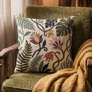 Puede incluir: Un cojín cuadrado con base de color crema y bordado en relieve tipo punch needle. El diseño muestra un patrón botánico de helechos, hojas de monstera y flores estilizadas en tonos verde salvia, verde oliva y rosa suave.