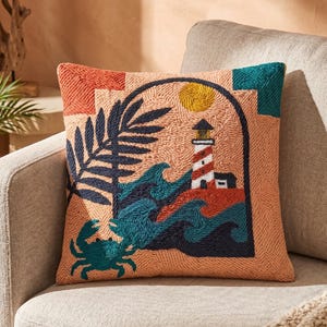 Puede incluir: Un cojín cuadrado con un diseño bordado texturizado sobre un fondo color melocotón. La escena muestra un faro rojo y blanco, olas de mar azul oscuro, una hoja de palmera grande, un sol amarillo y un cangrejo verde azulado dentro de un arco.
