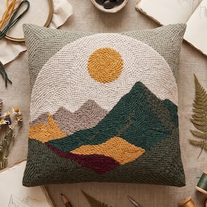 Könnte beinhalten: Ein quadratisches Kissen mit einem Punch-Needle-Landschaftsmotiv, das eine gelbe Sonne, dunkelgrüne Berge und einen strukturierten beigen Himmel zeigt. Das Kissen ist von Stickzubehör umgeben, darunter ein Holzrahmen, Garn und Skizzenbücher.