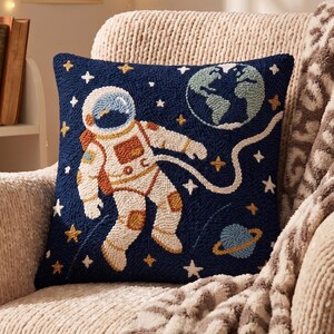 Könnte beinhalten: Ein quadratisches Zierkissen mit dunkelblauem Hintergrund und einem strukturierten Punch-Needle-Design eines im Weltraum schwebenden Astronauten. Das Motiv zeigt eine weiße Leine, eine hellblaue Erde, einen kleinen Planeten mit Ring und Sterne.