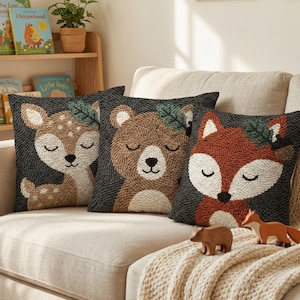 May include: Three square throw pillows featuring textured, punch-needle style illustrations of a deer, a bear, and a fox against a dark gray background. Each animal has closed eyes and a small green leaf accent on its head, creating a cohesive set.