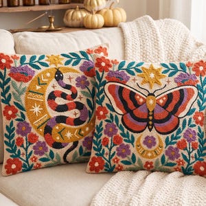 May include: Two square throw pillows featuring intricate punch needle embroidery on a cream background. One pillow displays a coiled snake and a crescent moon, while the other features a large butterfly, both surrounded by colorful floral motifs.