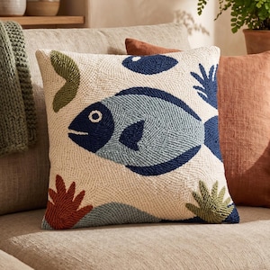 May include: A square throw cushion with a cream-coloured base featuring a textured punch needle design of a large blue fish in the centre. The pattern includes smaller abstract shapes in dark blue, olive green, and terracotta red colours.